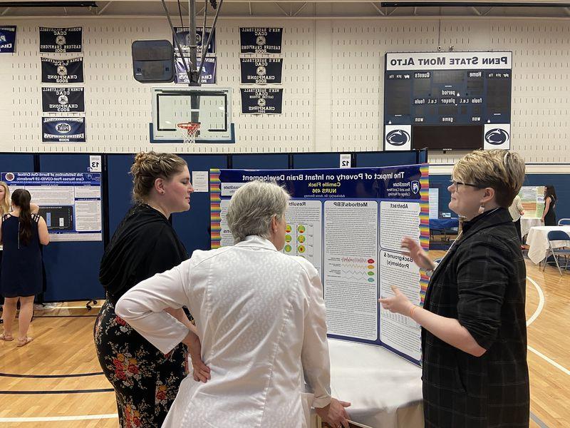 A student talks to others in front of a tri-fold board that reads "The Impact of Poverty on Infant Brain Development"