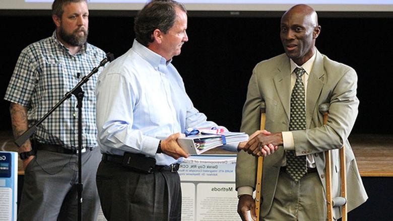 Chancellor Francis Achampong hands the WCHS project policies and procedures book to WCHS President Greg Duffey.