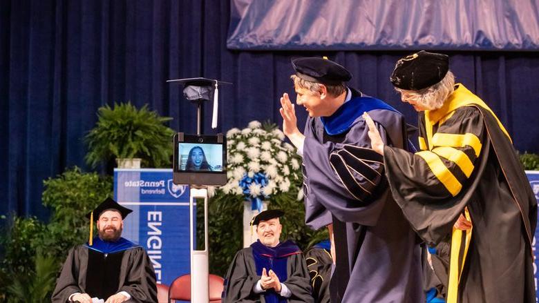 Two Penn State Behrend faculty members wave to a student whose face is on the monitor of a robot.