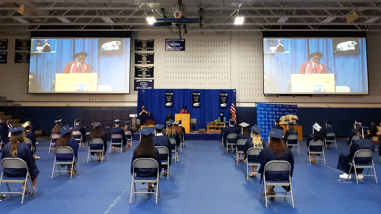 Students in regalia in the campus gym listening to remarks from the chancellor